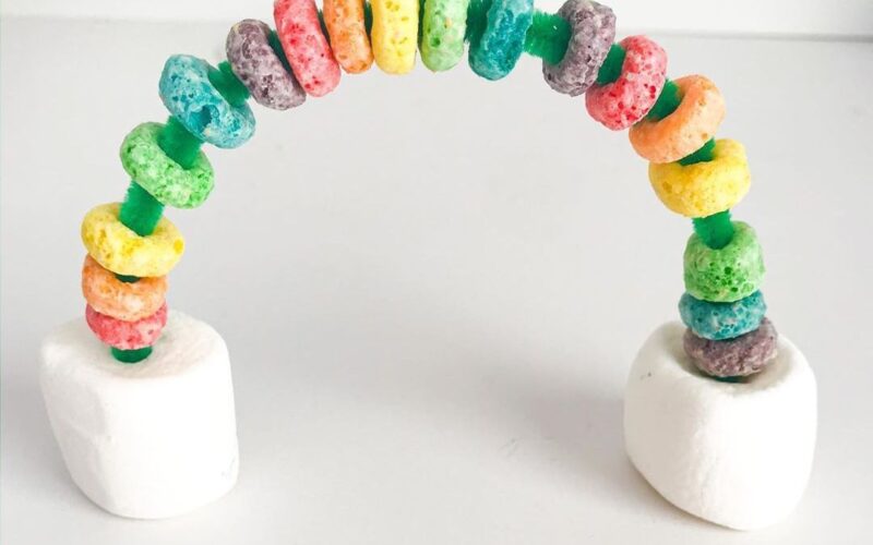 Two marshmallows serve as the base for a rainbow that is made from a curved pipe cleaner with froot loops strung on it.