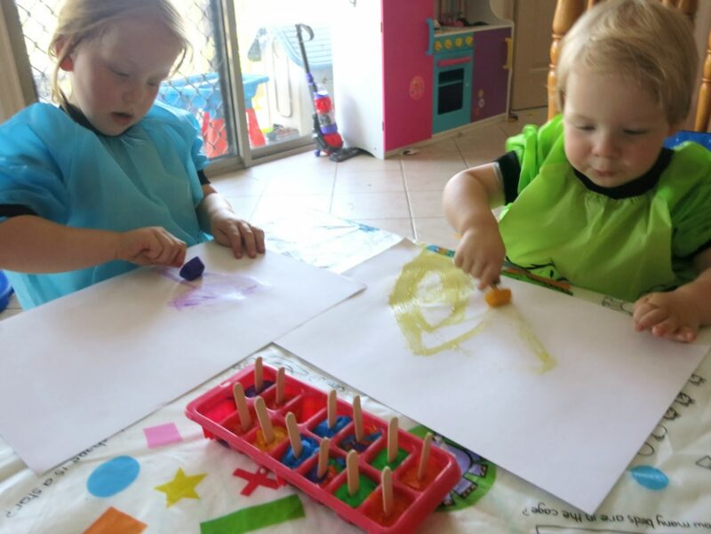 Two small children are seen painting. An ice cube tray with paint and popsicle sticks coming out is also shown.