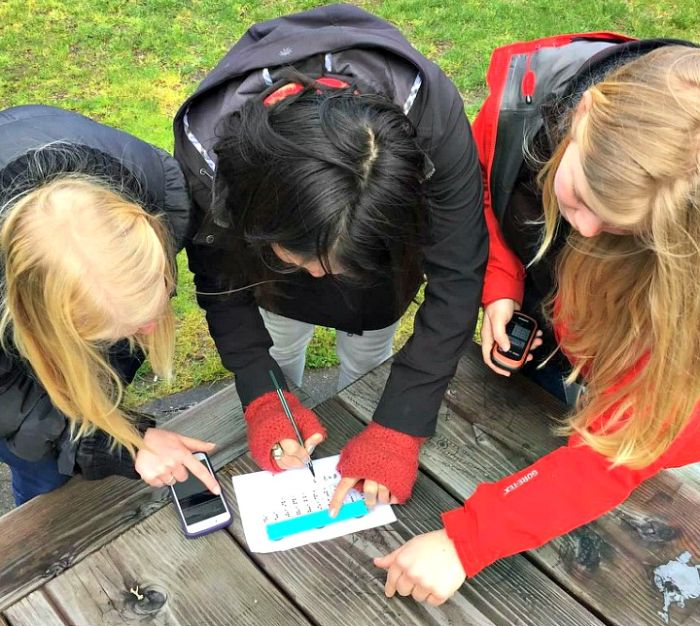 A group of students working together to solve a puzzle on a picnic table, as they look for a geocache together