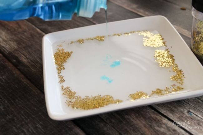Flat white dish filled with water with gold glitter on top, being scattered by a drop of dish soap (Germ Science Experiments)