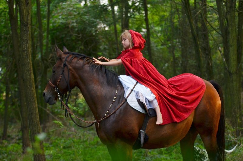 A young girl wearing a red hooded cape riding a brown horse in the forest