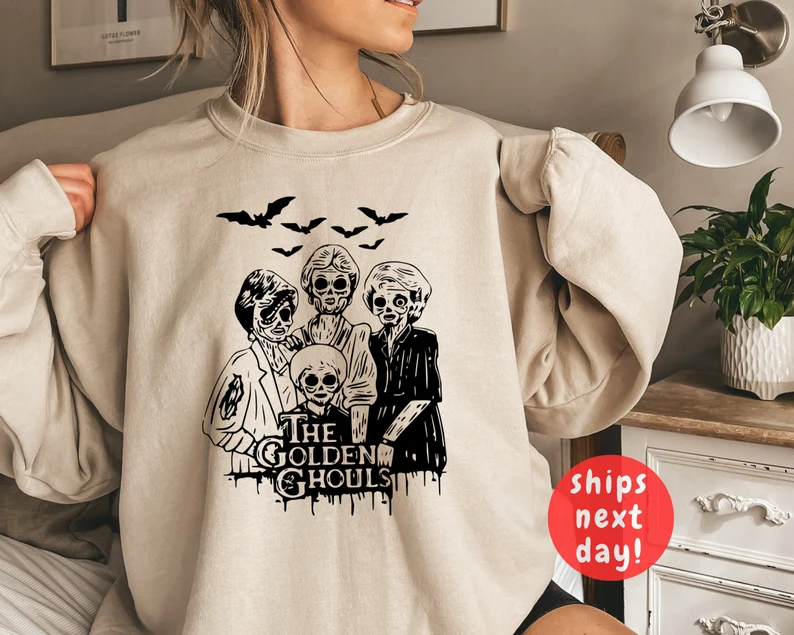 A woman is shown from the neck down wearing a beige sweatshirt that says Golden Ghouls and features the Golden Girls with their faces turned into ghouls.