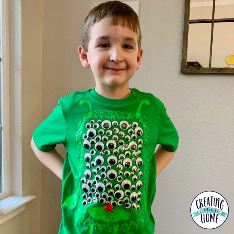 A little boy wears a green shirt that has googly eyes all over it (100th day of school shirt ideas)