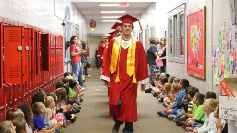 High school seniors in graduation gowns parade down a school hallway lined with elementary students
