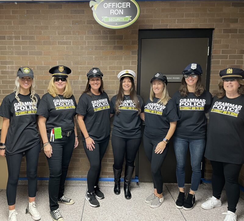 Seven women are shown wearing police officer hats and wearing black shirts that say Grammar Police.