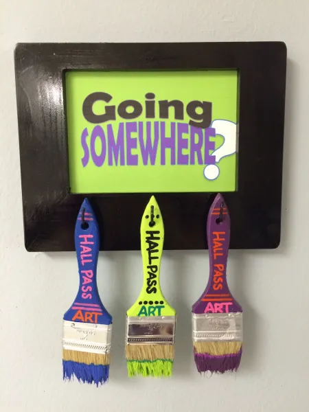a painted sign with three paintbrushes hanging from it for use as hall passes for classroom procedures 