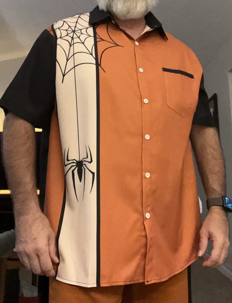 A man is shown from the neck down wearing a button up shirt in orange, black, and beige. A spider web is on the shoulder and a spider is crawling up the shirt.