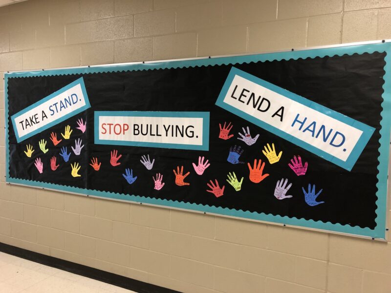 October bulletin board ideas includes this one on a black background. Multi-colored hands cover the board. The text reads "take a stand," "stop bullying," and "lend a hand." 