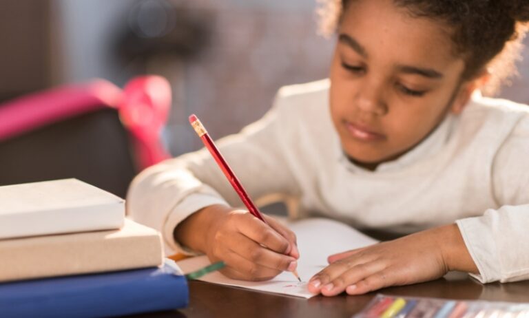 african american child writing in a notebook at a desk with stacks of book