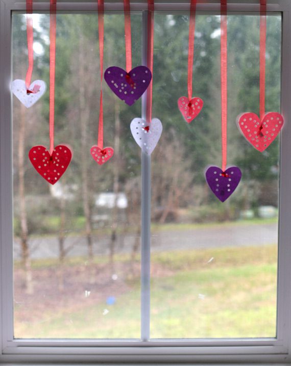 white, purple, pink, and red hearts are hanging from strings in front of a window. There are holes punched in the hearts (Valentine's Day Crafts for Preschoolers)