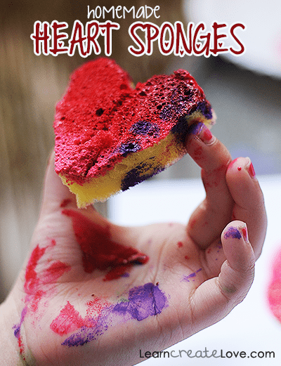 A hand is shown covered in red and purple paint and is holding a sponge that has been cut into a heart shape to make a stamp.