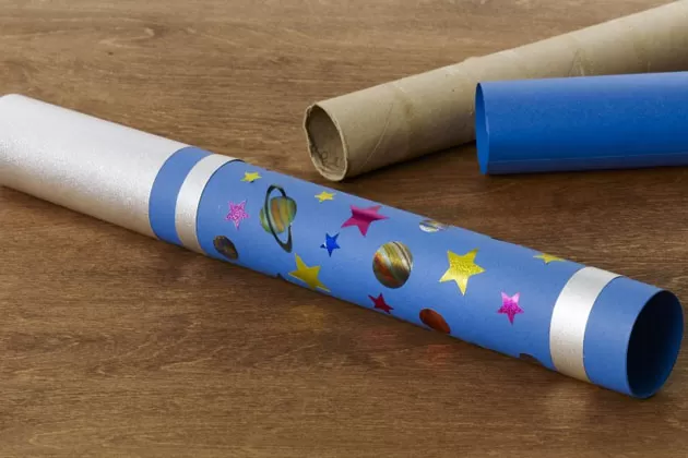 A homemade telescope is made from a cardboard roll that is decorated with blue and silver cardstock and space themed stickers.