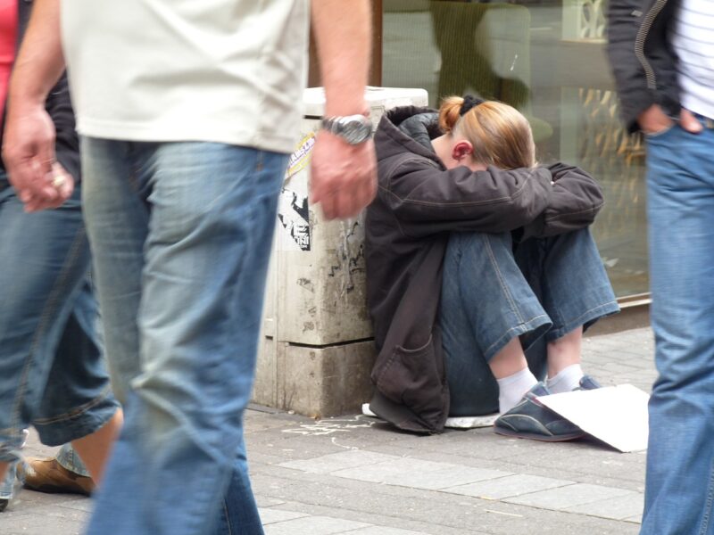 Woman sitting on a sidewalk with her head on her knees as others walk by