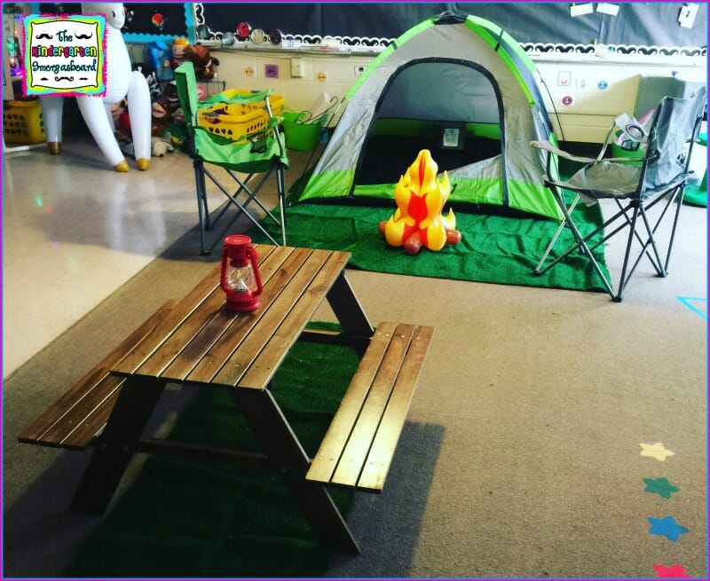 A classroom is shown that has been setup to look like a campsite. A picnic table, a green tent, and an inflatable campfire are shown. Camping Classroom Theme