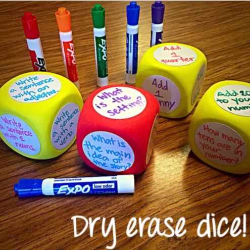 Several dry erase markers are shown in front of dice with things written on them with said markers.