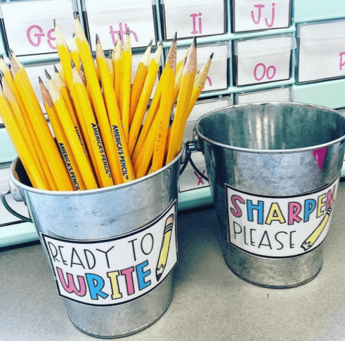 Two metal buckets have signs on them and one is filled with pencils. The one filled with pencils says Ready to Write and the second bucket says sharpen please.