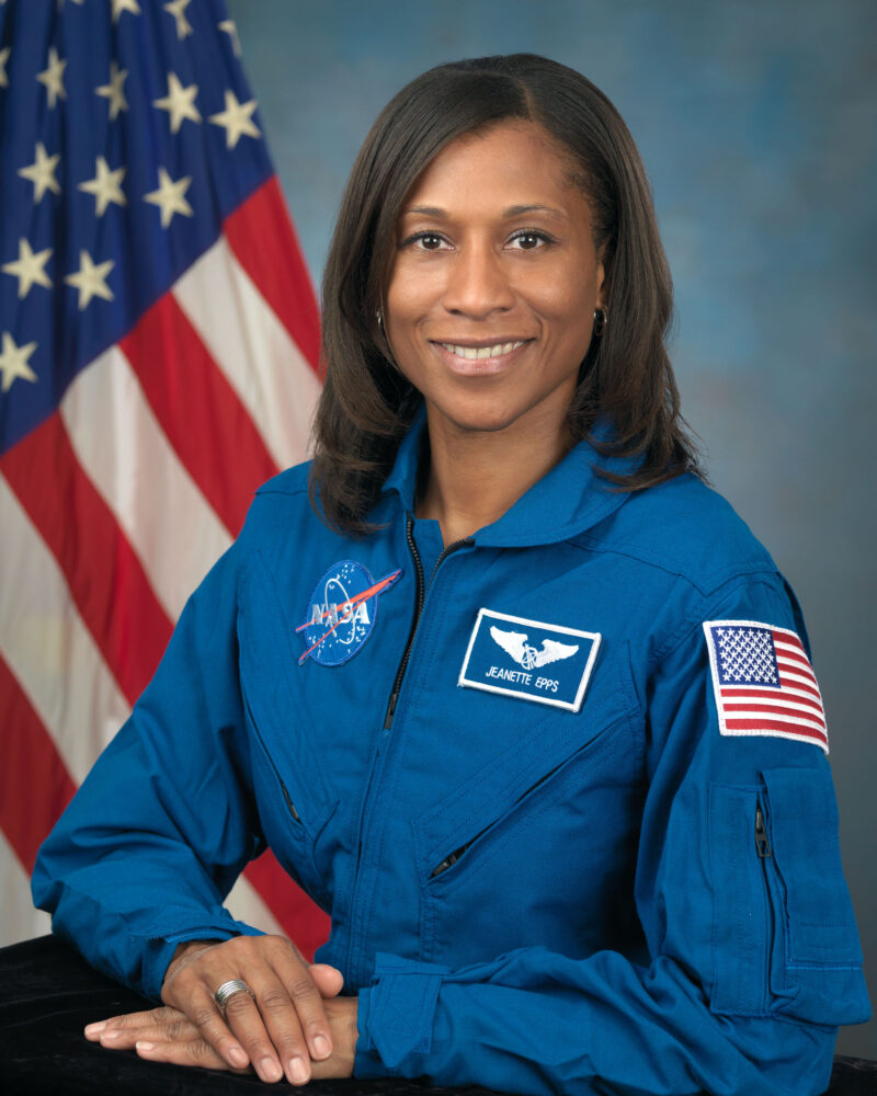A woman wearing a NASA suit is seen smiling in front of an American flag.