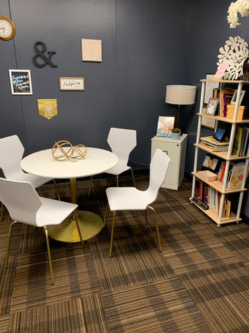 a collaborative workspace with a white table and 4 white chairs and a bookcase filled with books and accessories.