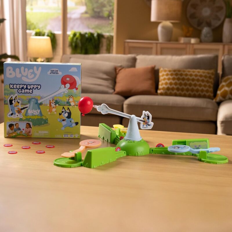 best board games for preschoolers include this game. A box is shown with the dog cartoon character Bluey on it. A contraption is on a table and a fake balloon is suspended on a see-saw.