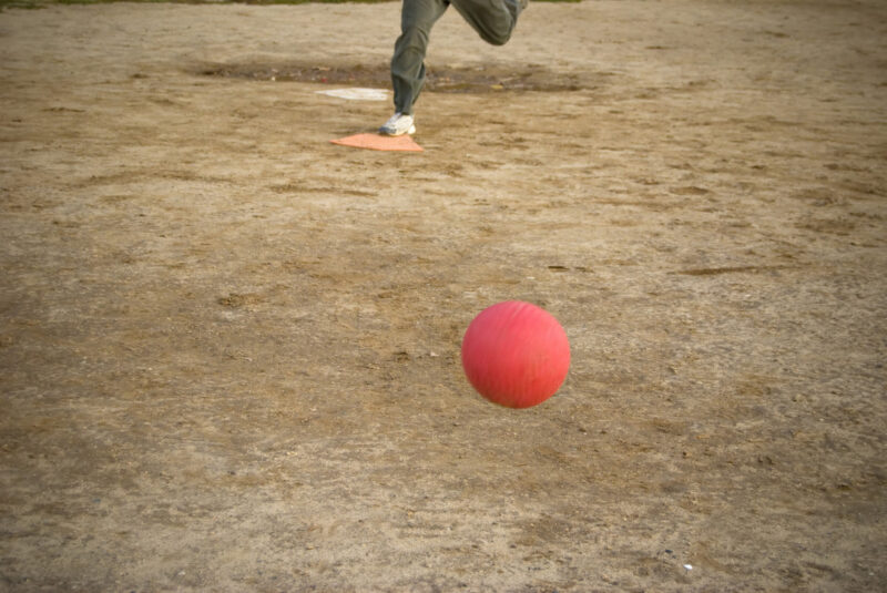 A red kickball rolls toward home plate after it is pitch - the person pictured here on plate is approaching the kick.