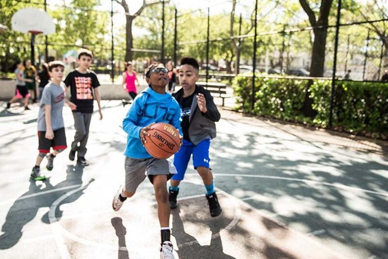 A boy is seen holding a basketball while another plays defense. More kids are seen in the background on the basketball court.