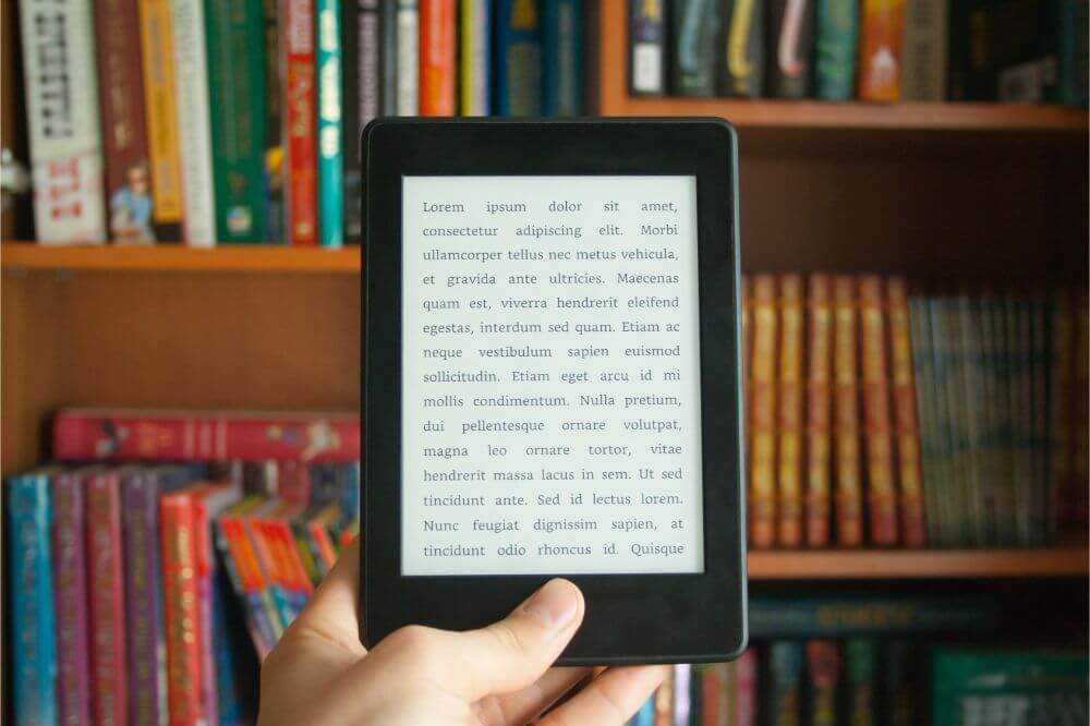 A hand is shown holding a kindle device up in front of a bookshelf of actual books (compare and contrast essay example)