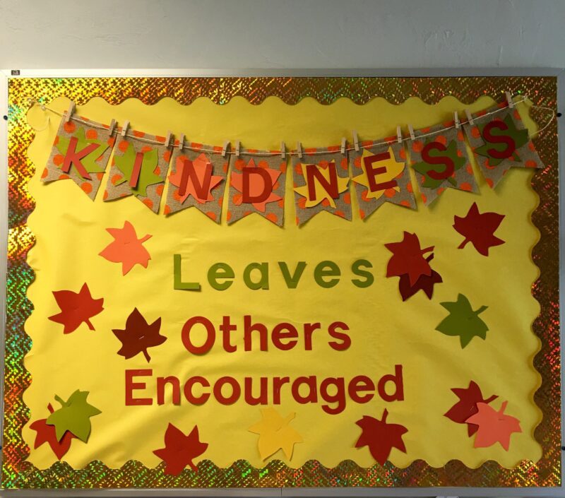 A yellow bulletin board reads Kindness leaves others encouraged. Kindness is in a banner across the top and fall leaves are sprinkled throughout.