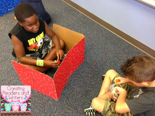 two kids building with legos, with one of them sitting behind a barrier to hide his creation 