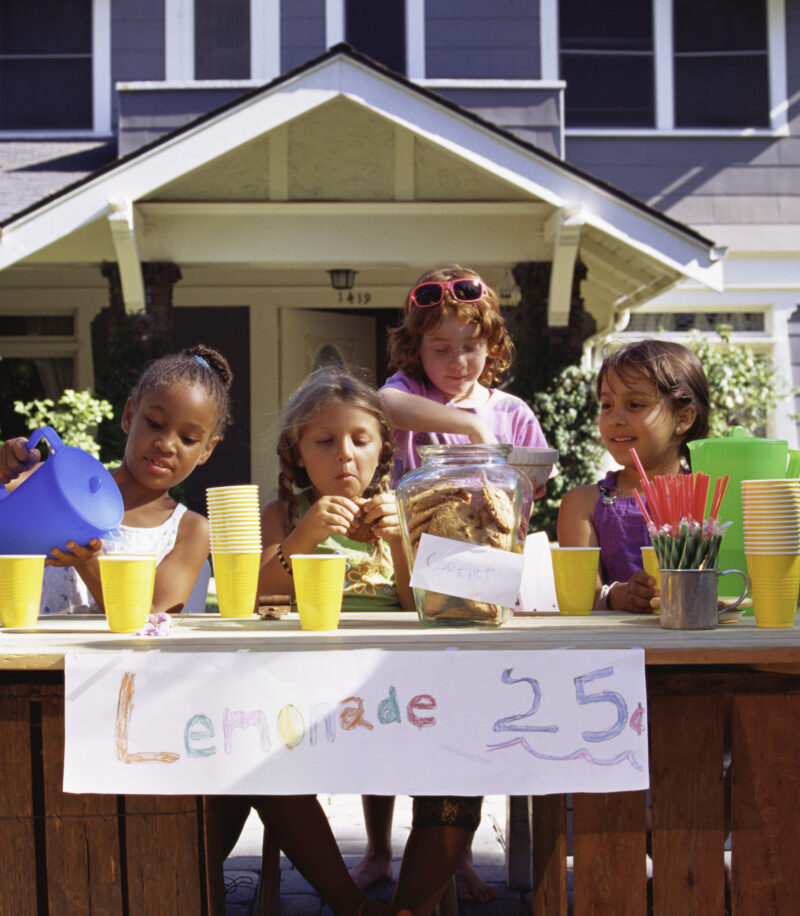 Girls with lemonade stand in front of house.