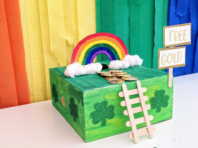 A leprechaun trap is created from a box painted green with a Popsicle stick ladder and clouds and a rainbow on top. 