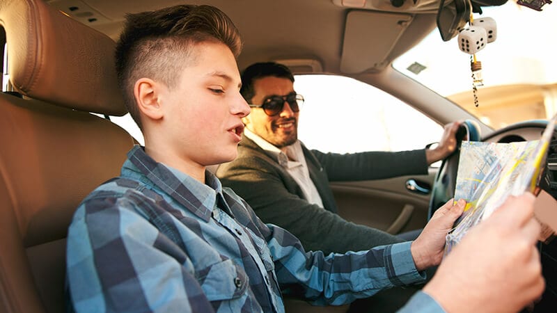Dad driving car and son reading a map, as an example of life skills for teens.
