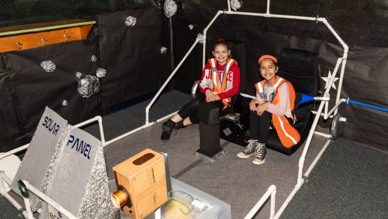 Two girls sit in a large lunar rover model that has been constructed from PVC piping.- space-themed classroom