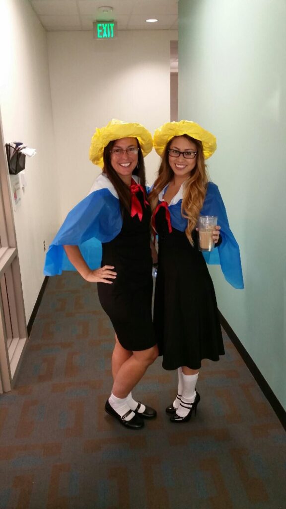 Two women are seen wearing matching outfits. They are wearing yellow hats, navy dresses, red neckerchiefs, and blue capes.