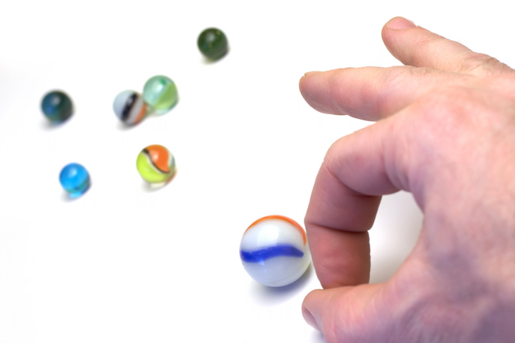 A hand is seen flicking a marble toward other marbles.