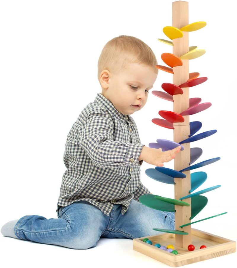 A toddler boy is sitting in front of a tall wooden tree with brightly colored leaves. The base is holding marbles in this example of marble games.