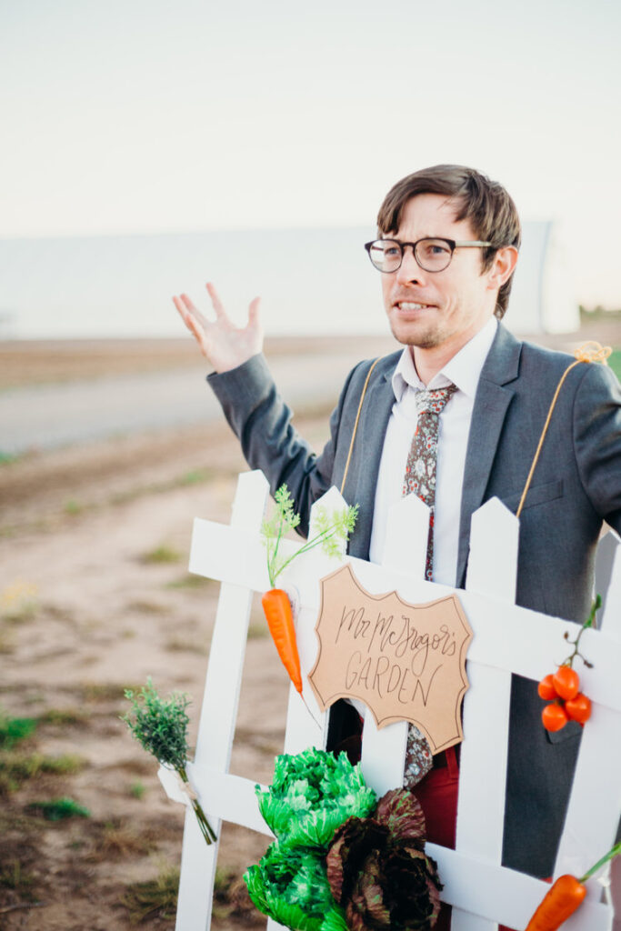 A man in a suit has a white picket fence hanging off of him and vegetables.- book character costume