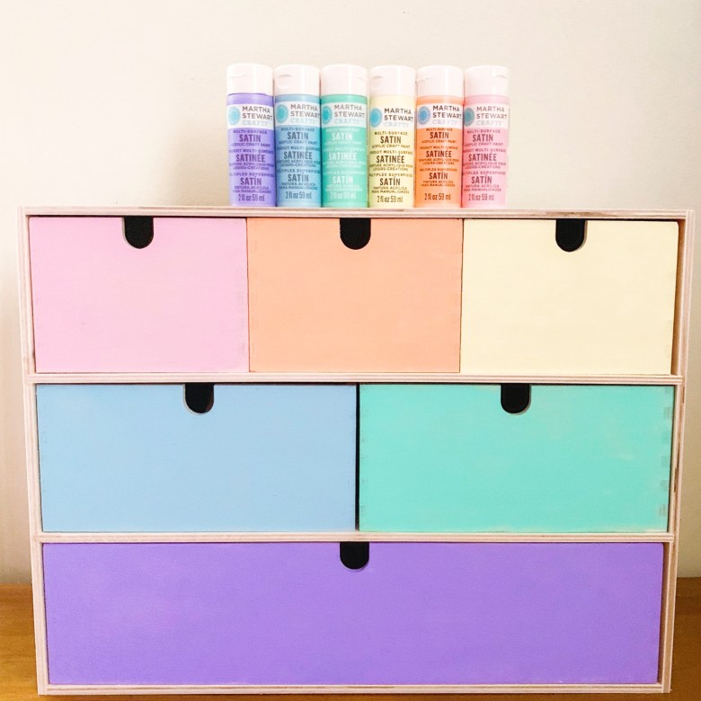 A chest of draws is painted in pastel colors with tubes of paint sitting on the top of it.