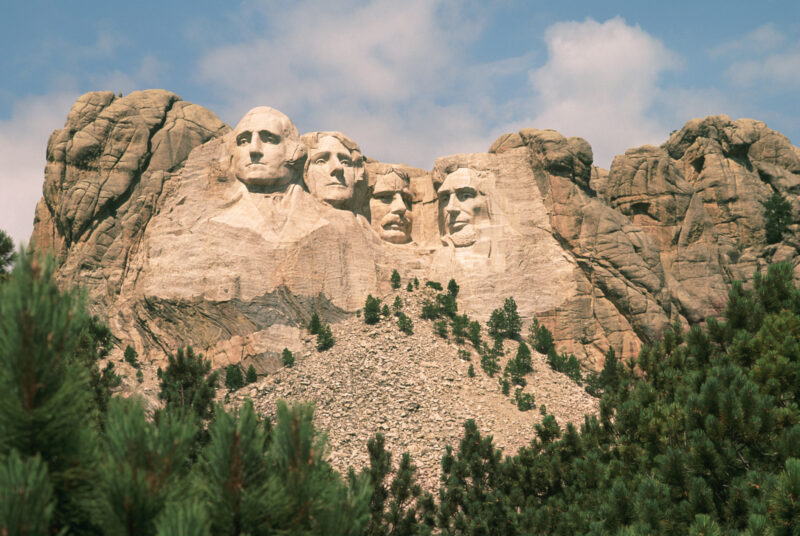 View of Mount Rushmore. The carved faces of the four historical figures, George Washington, Thomas Jefferson, Theodore Roosevelt and Abraham Lincoln are framed by a brilliant blue sky and puffy white clouds. Bright green coniferous trees in the foreground provide contrast to the granite stone into which the faces are carved