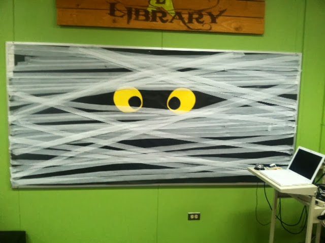 A library sign hangs above a board that is black with yellow eyes peeking out and white crepe paper wrapped around it to look like a mummy.