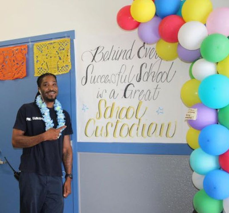 School custodian standing by a sign and balloons celebrating National Custodian Day