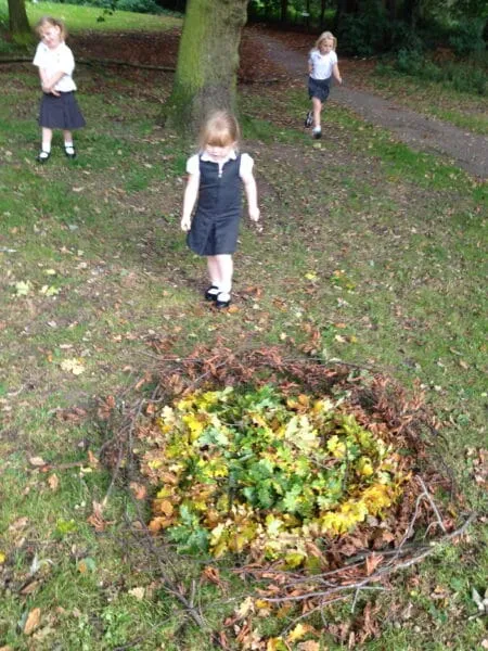 Several little girls stand behind a circle of leaves and sticks that have been assembled into a collaborative art project.