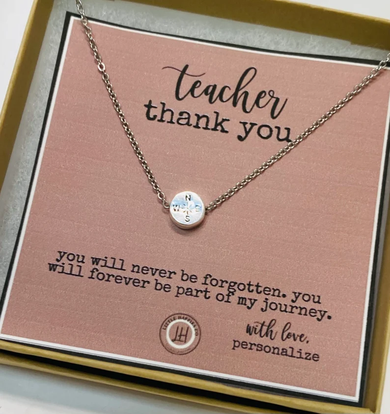 A silver necklace with a compass on it is on a cardboard backing that is also a thank you note (personalized teacher gifts)