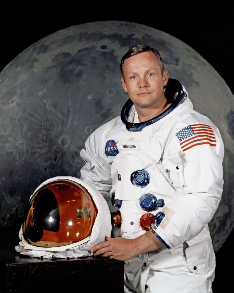 A young man is in a full astronaut outfit holding his helmet. Famous engineers include Neil Armstrong who is posing in front of the moon. 