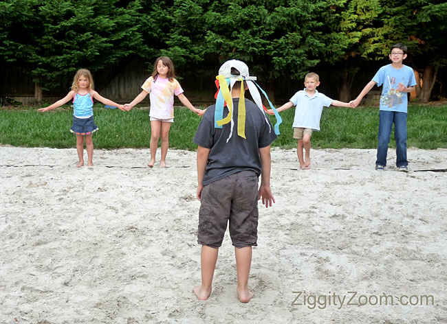 Children are seen in the background holding hands. One child has their back to the camera facing the other children (tag games)