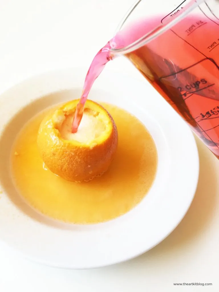 An orange is shown in a bowl with a pitcher of red liquid pouring into the cut off top of it.