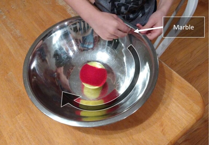 A metal mixing bowl has a tennis ball inside it and a child's hand is holding a marble at the top.