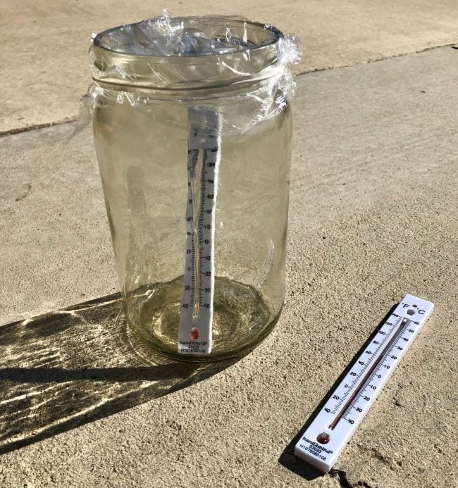 Glass jar covered in plastic wrap with a thermometer inside, next to a thermometer lying on the ground 