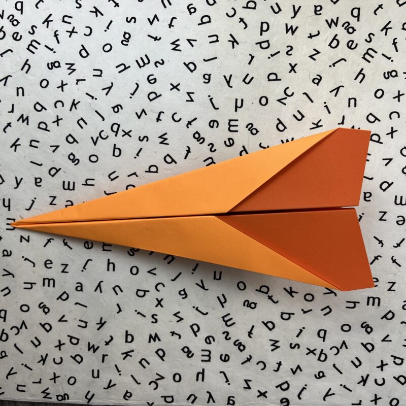 Finished orange dart paper airplane on black and white alphabet background.