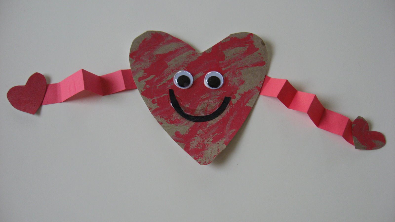 A cardboard heart has a smiley face and googly eyes on it. The arms are made from folded construction paper. 