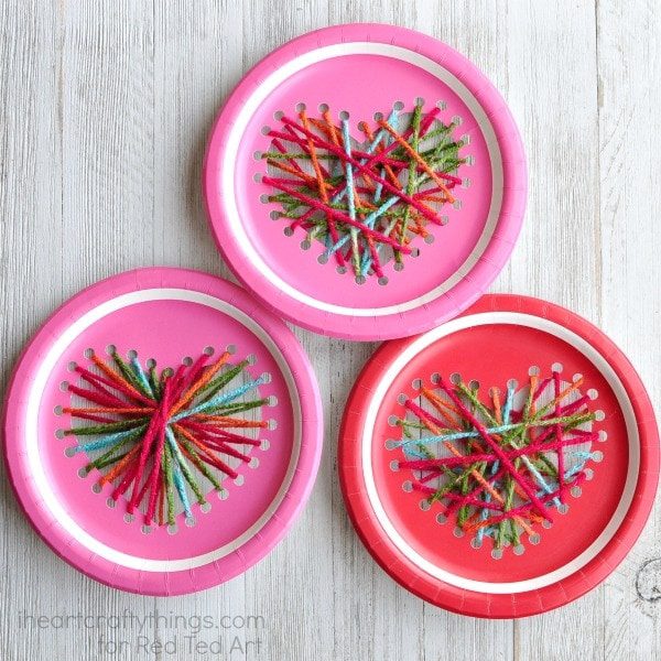 Three pink and red paper plates are shown with hole punches in the shape of hearts on them. They have yarn strung between the holes.